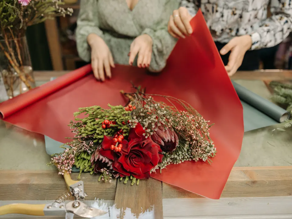 Twee bloemisten rollen rood inpakpapier uit over een boeket van rode rozen en groen op een houten werktafel, met een snoeischaar ervoor