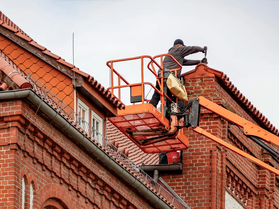 Dakwerker op een hoogwerker bij een rood bakstenen gebouw met pannendak en dakgoot