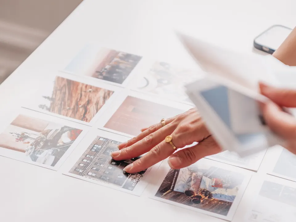 Een hand legt afgedrukte foto’s op een witte tafel uit, met een telefoon op de achtergrond