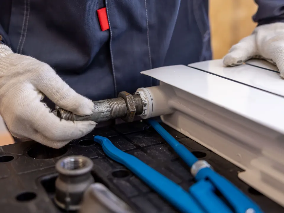 Handen met werkhandschoenen monteren een leiding aan een radiator, met een blauwe waterpomptang ernaast