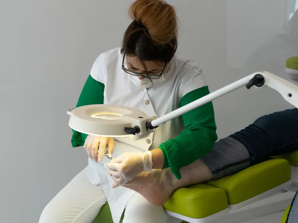 Voetverzorger met mondkapje en handschoenen behandelt een voet in een behandelstoel onder een loeplamp
