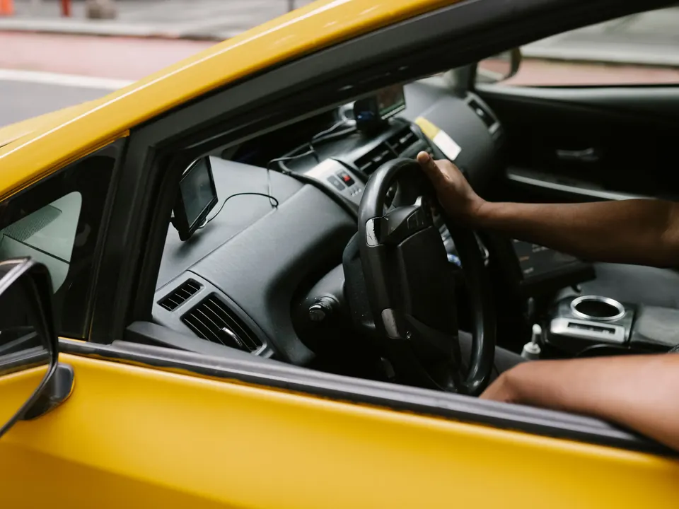 Chauffeur met een hand aan het stuur van een gele taxi, gezien door het zijraam