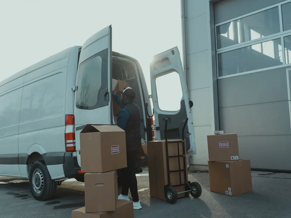 Bezorger laadt kartonnen dozen in een witte bestelbus bij een magazijn, steekwagen ernaast
