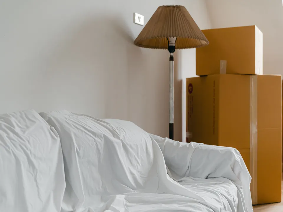 Afgedekte bank en gestapelde verhuisdozen naast een staande lamp in een lichte kamer