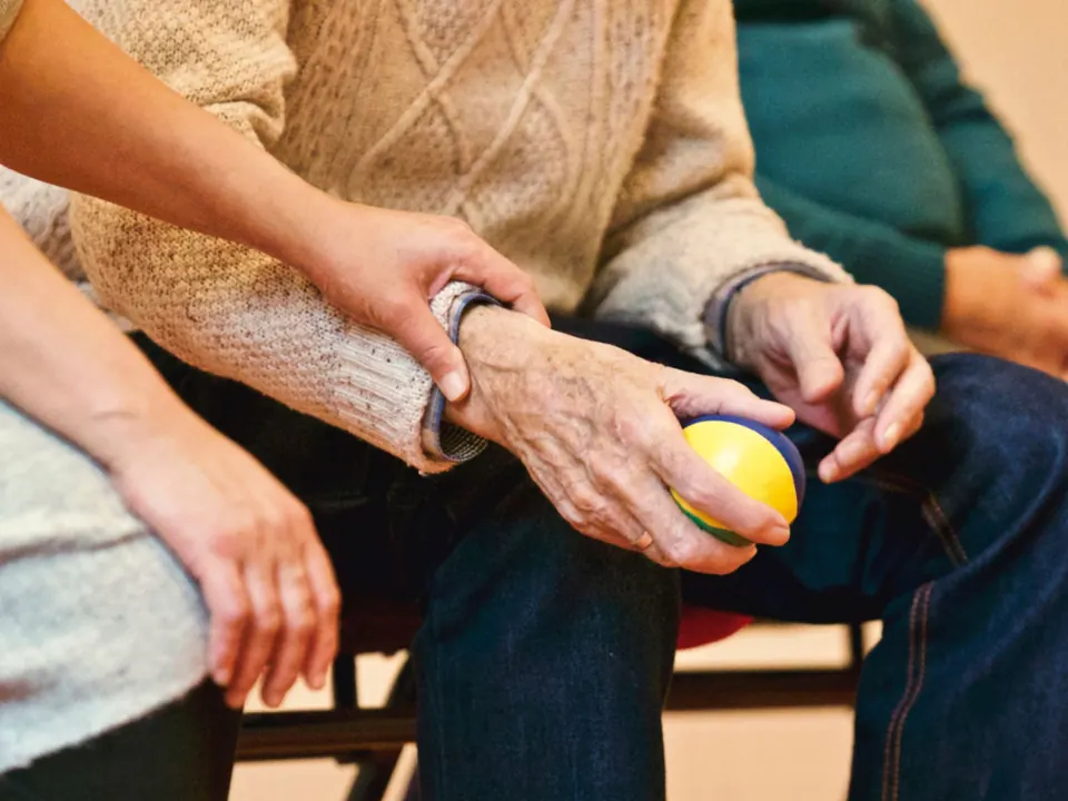 Een zorgverlener houdt de arm van een oudere cliënt vast die een zachte bal in de hand heeft